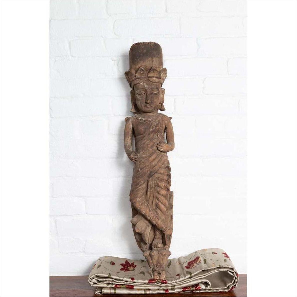 Brown wood statue of Hindu deity, hand carved from Gujarat, displayed on cloth against a white brick wall indoors.