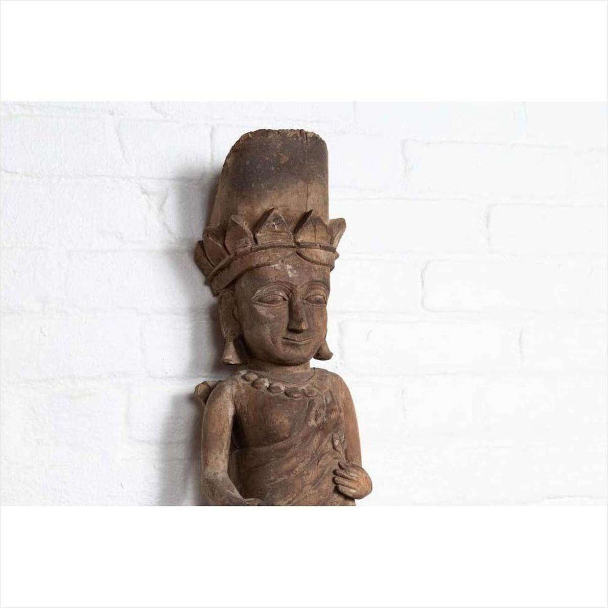 Brown hand-carved wooden statue of a Hindu deity with broken crown, set against white brick background.