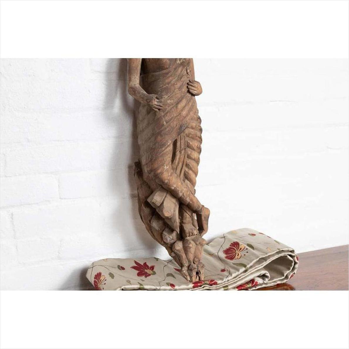 Brown wood carved statue of Hindu deity from Gujarat seated on floral fabric against a white wall indoors.