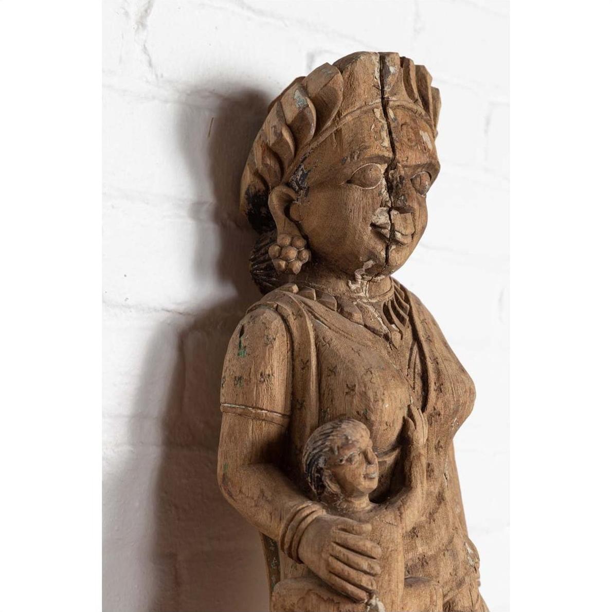 Brown wood statue of mother and child, hand carved in Indian temple style, set against a white brick wall indoors.