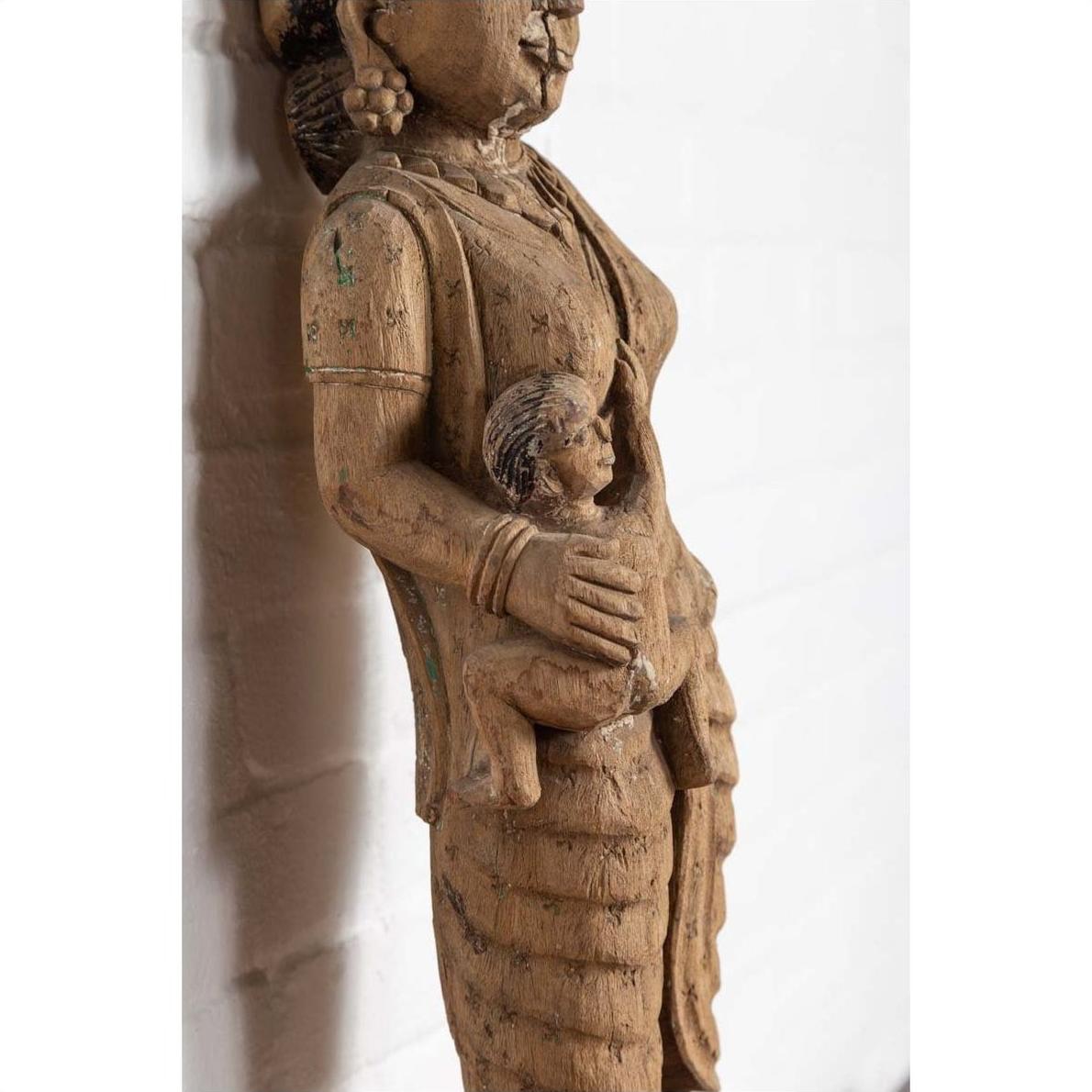 Brown wood mother and child statue, hand carved, Indian temple style, placed in front of a white brick wall indoors.