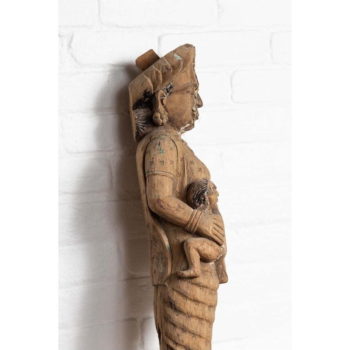 Brown wood mother and child temple carving statue, hand carved, side view, against white brick wall indoors.