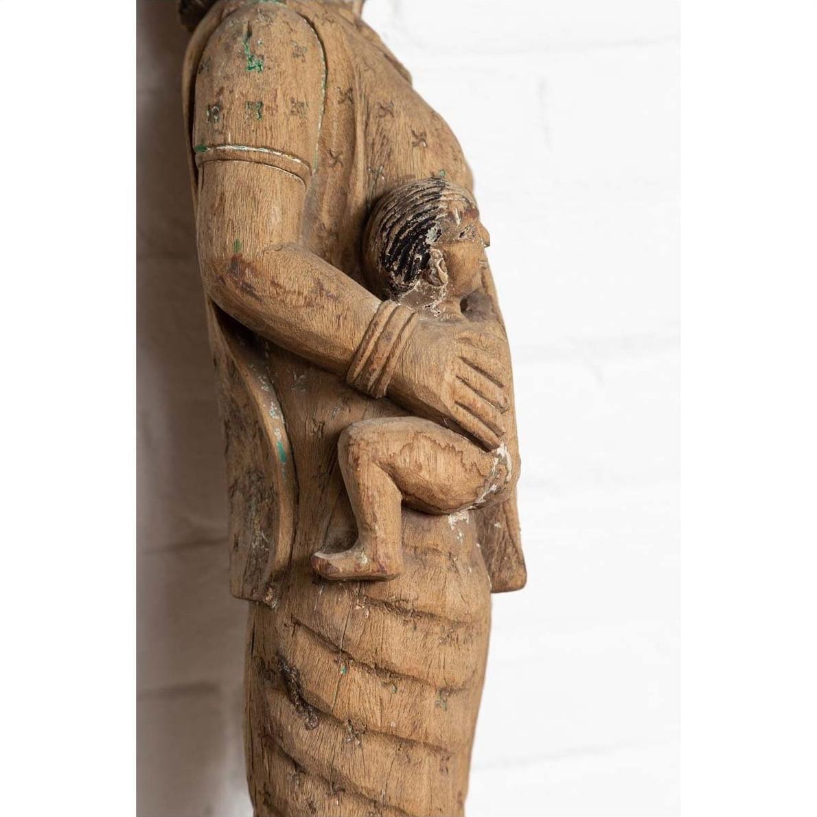 Brown hand-carved wood statue of a mother holding child, Indian temple carving from Gujarat, on white background.