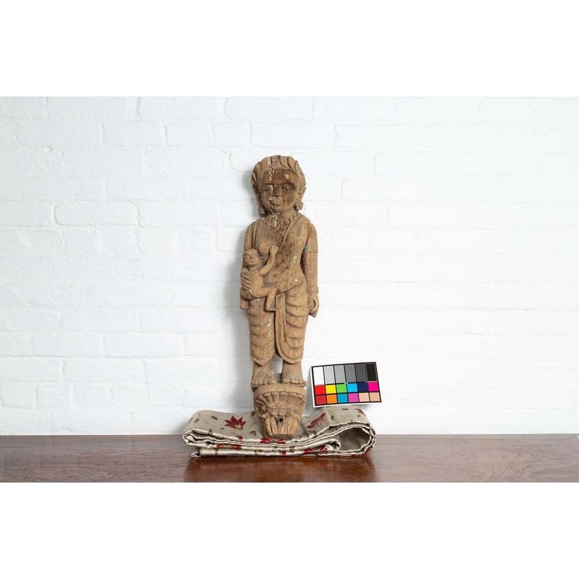 Brown hand-carved wood statue of mother and child from Gujarat, shown with color card against white brick background.