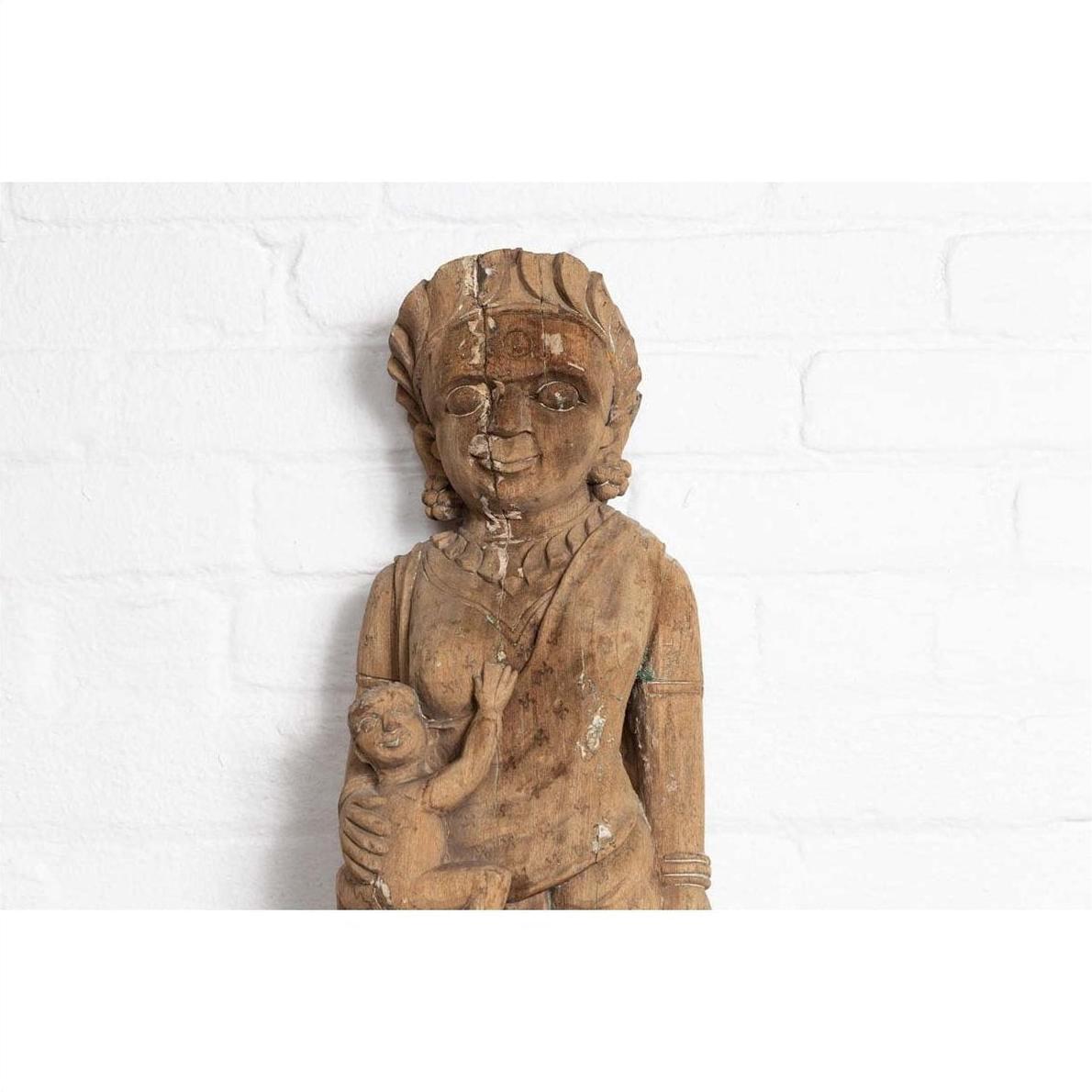 Brown wood mother and child statue, hand-carved with Indian temple design, placed in front of a white brick wall.
