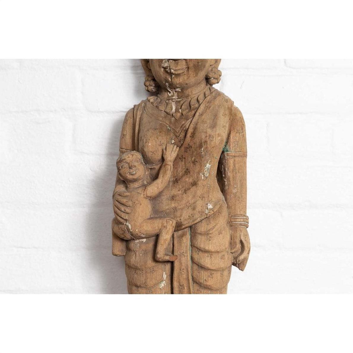 Brown hand-carved wooden temple statue of a mother holding her child, set against a white brick wall.