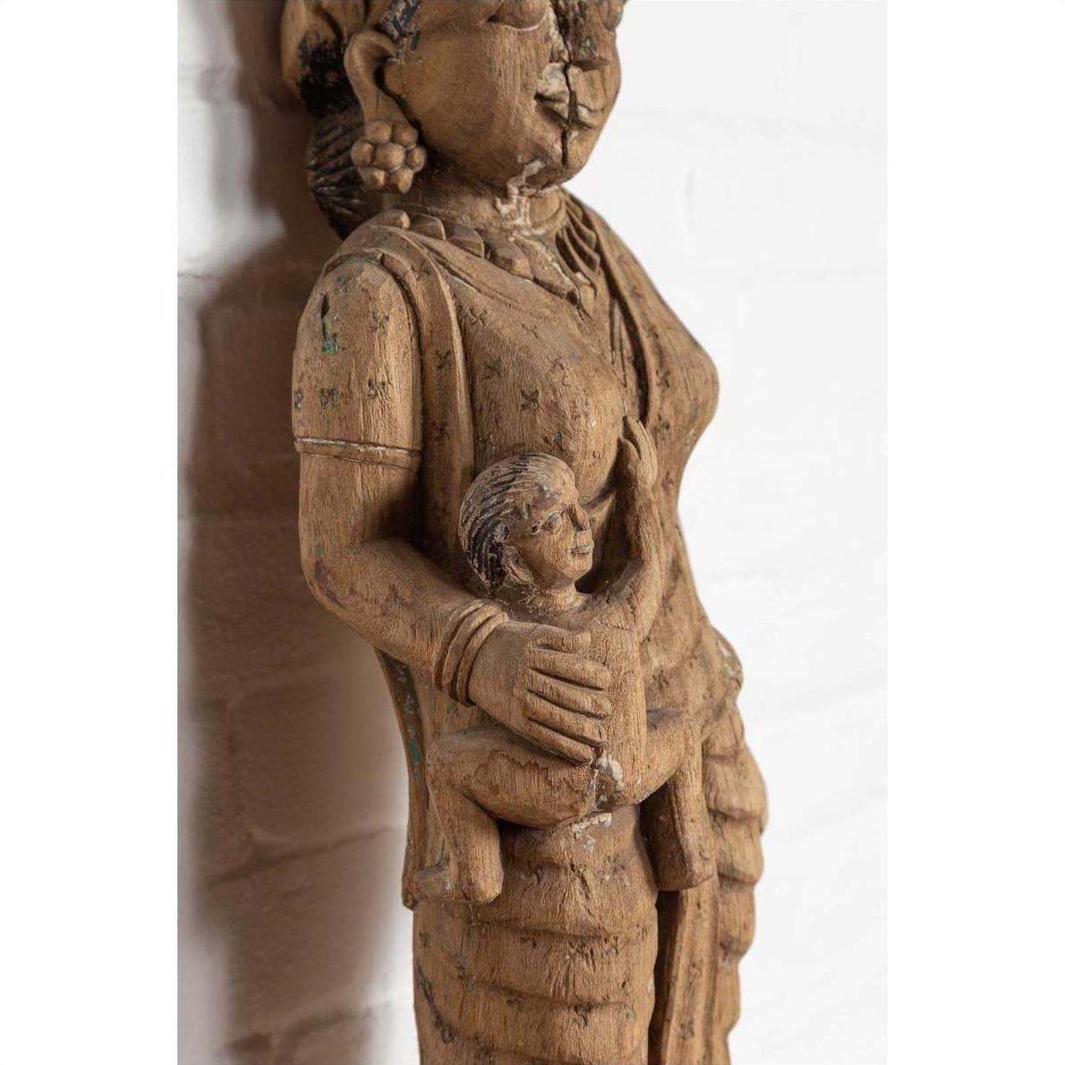 Brown wooden statue of mother and child, hand carved in Indian temple style, with white brick background.