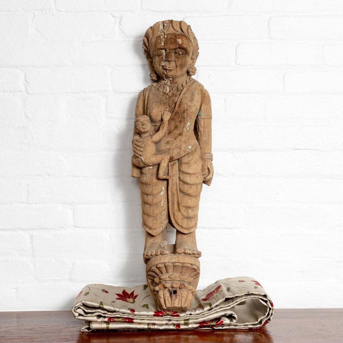 Brown wooden statue of mother and child with Indian temple carvings, displayed on patterned fabric against white brick wall.