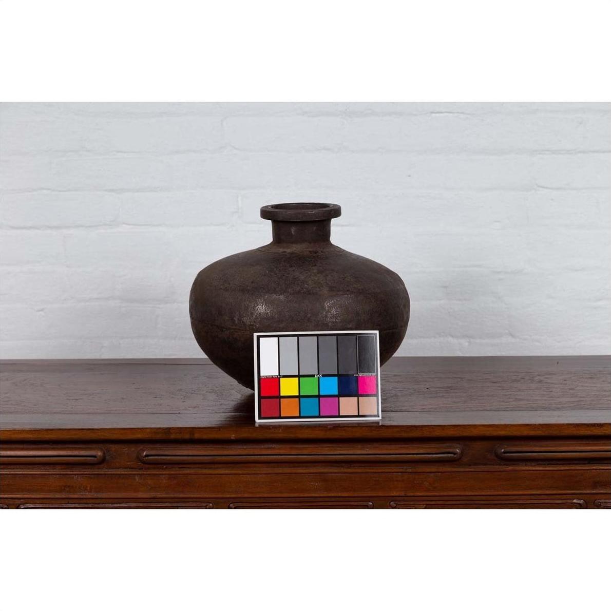 Brown metal Indian 19th c. water jar with wide belly and protruding lip on wood surface, color card in front.