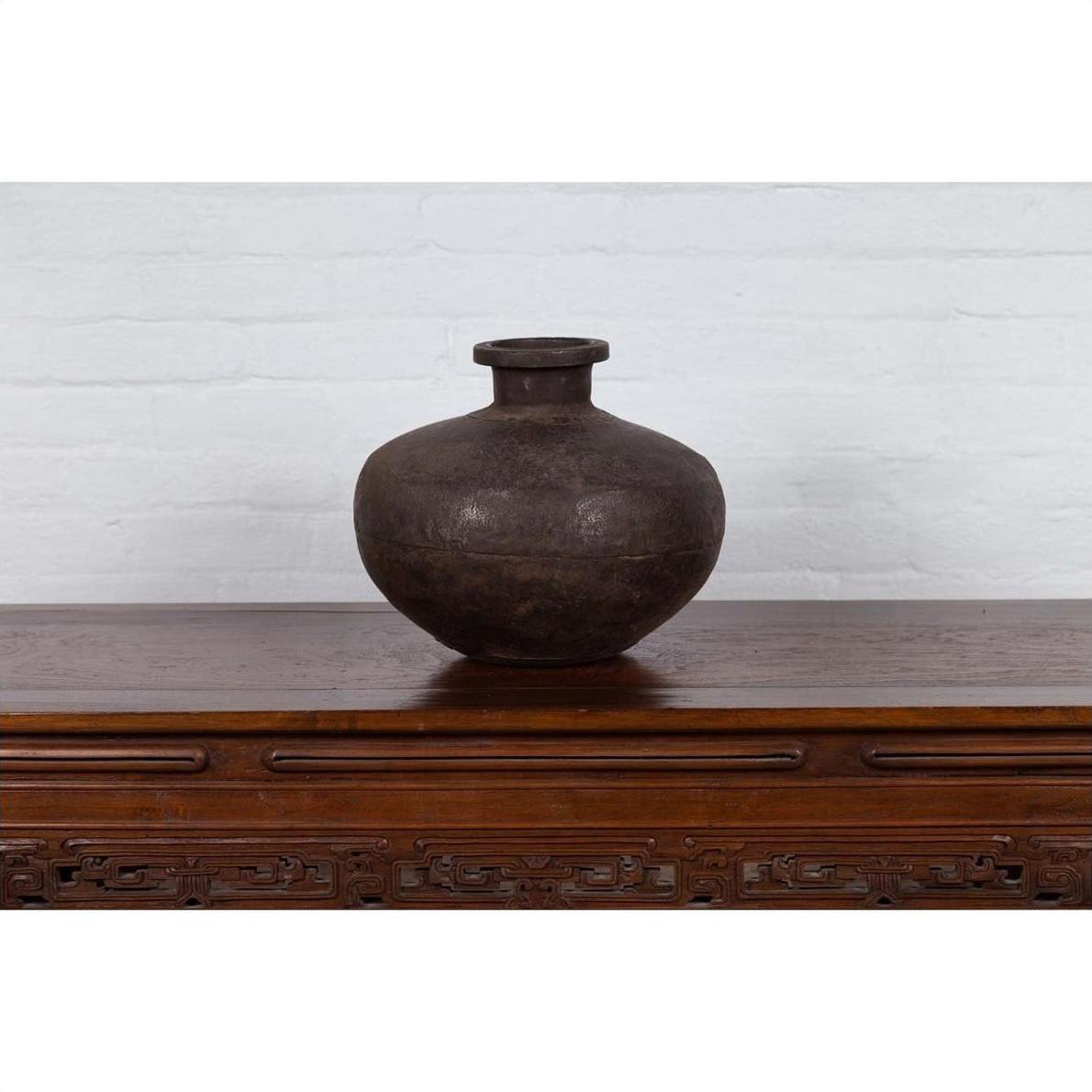 Dark clay vase and 19th century Indian metal water jar on carved table against white brick wall.