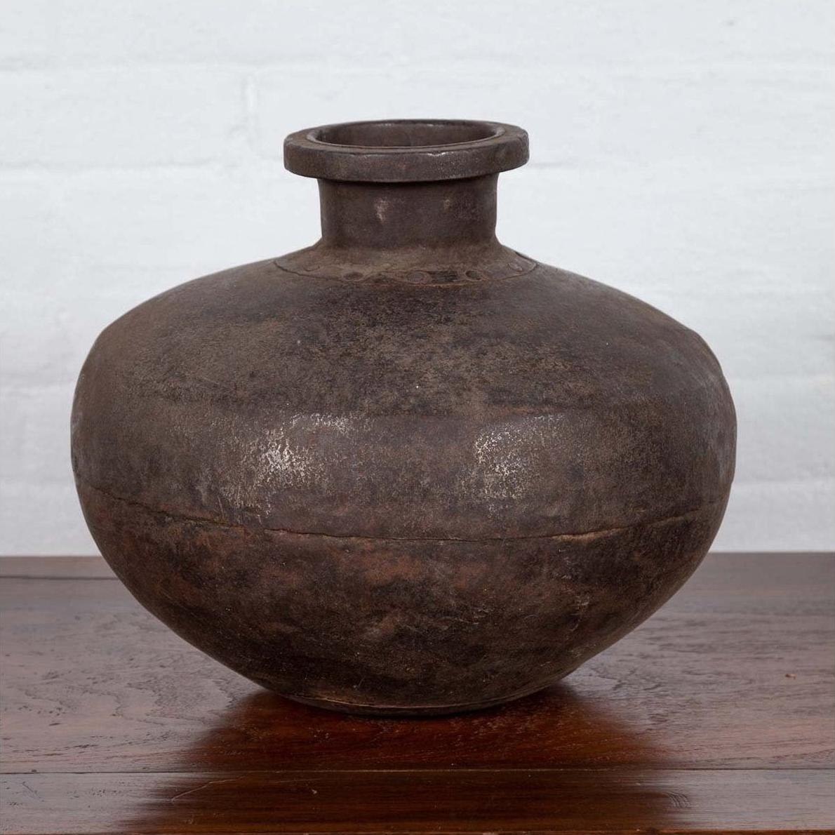 Metal Indian water jar with a generous belly and protruding lip on wooden surface near a white wall, 19th century.