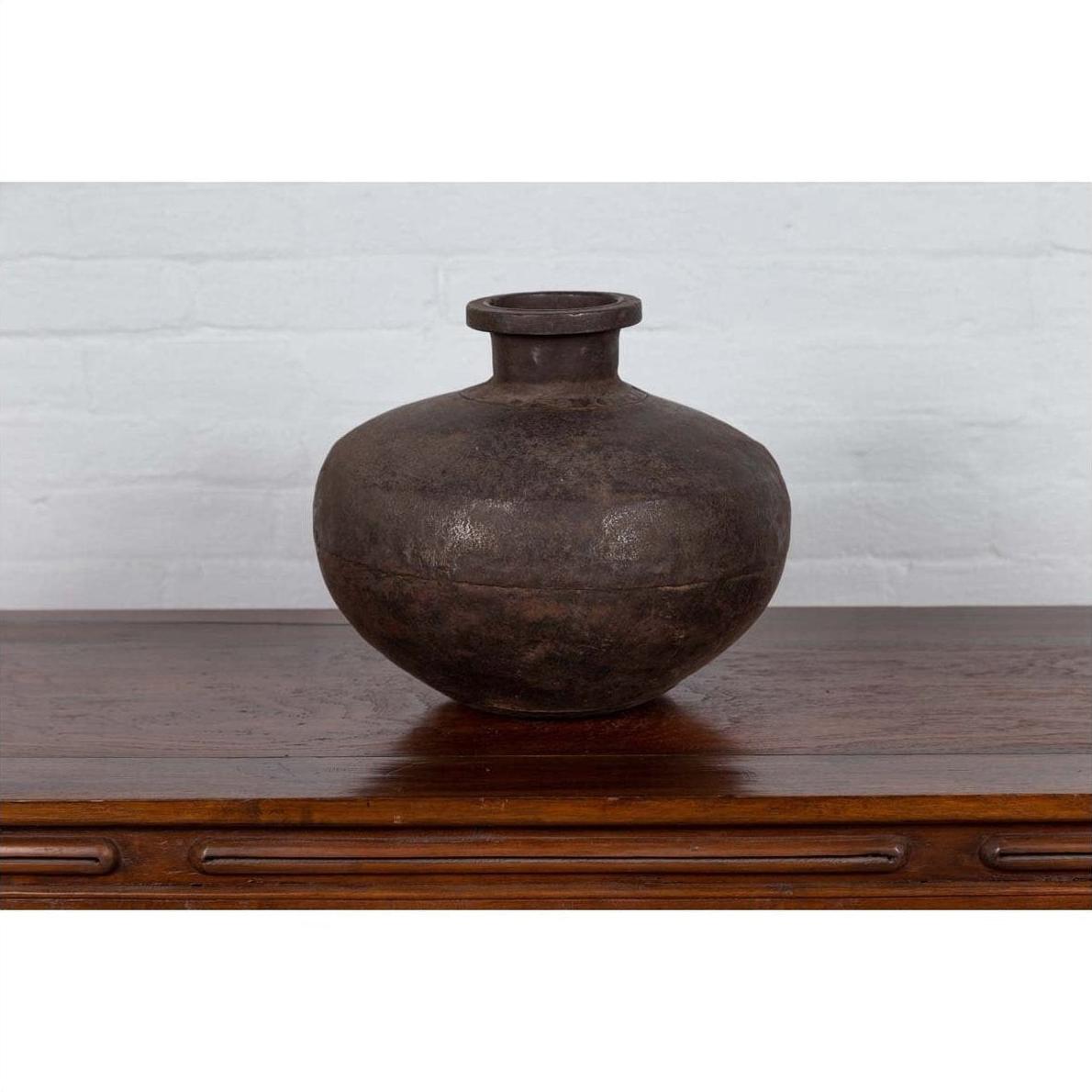 Bronze metal Indian 19th-century water jar with rounded belly and protruding lip on wood surface, white brick wall background.