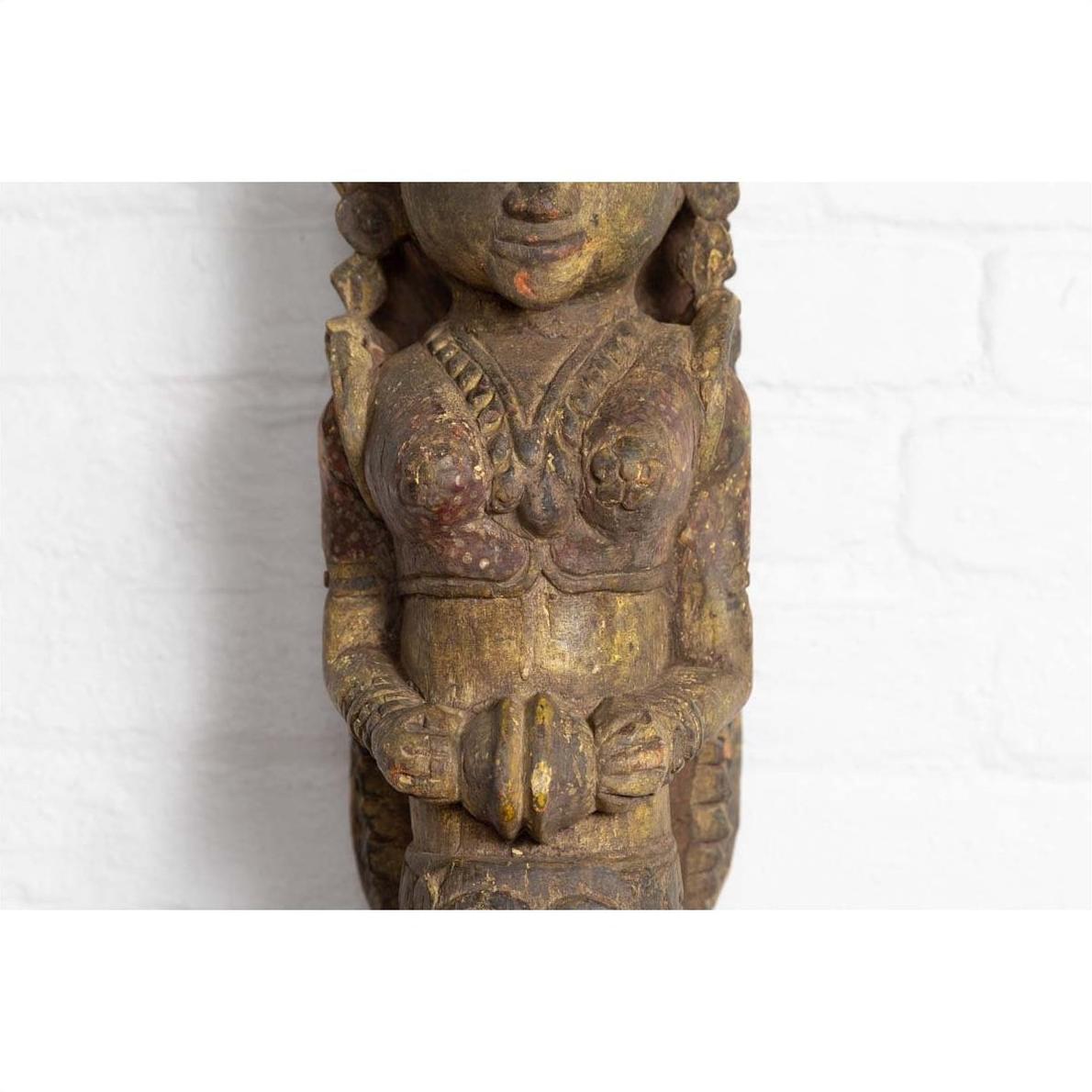 Close-up of polychrome hand carved wooden statue of Indian celestial musician against a white brick wall background.