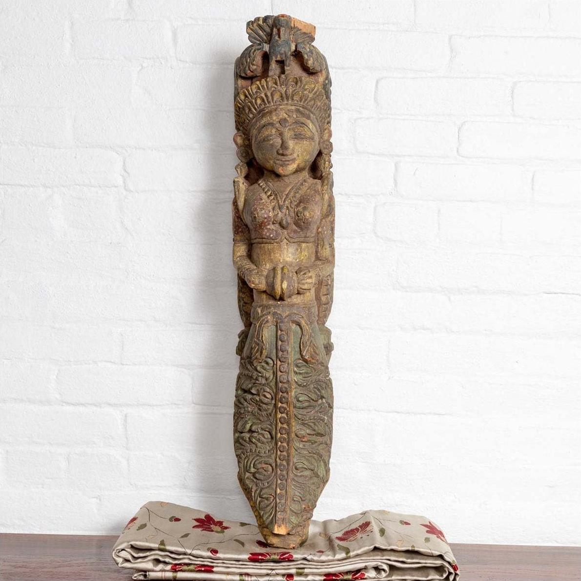 Polychrome hand carved wooden statue of an Indian celestial musician on cloth with a white brick wall background.