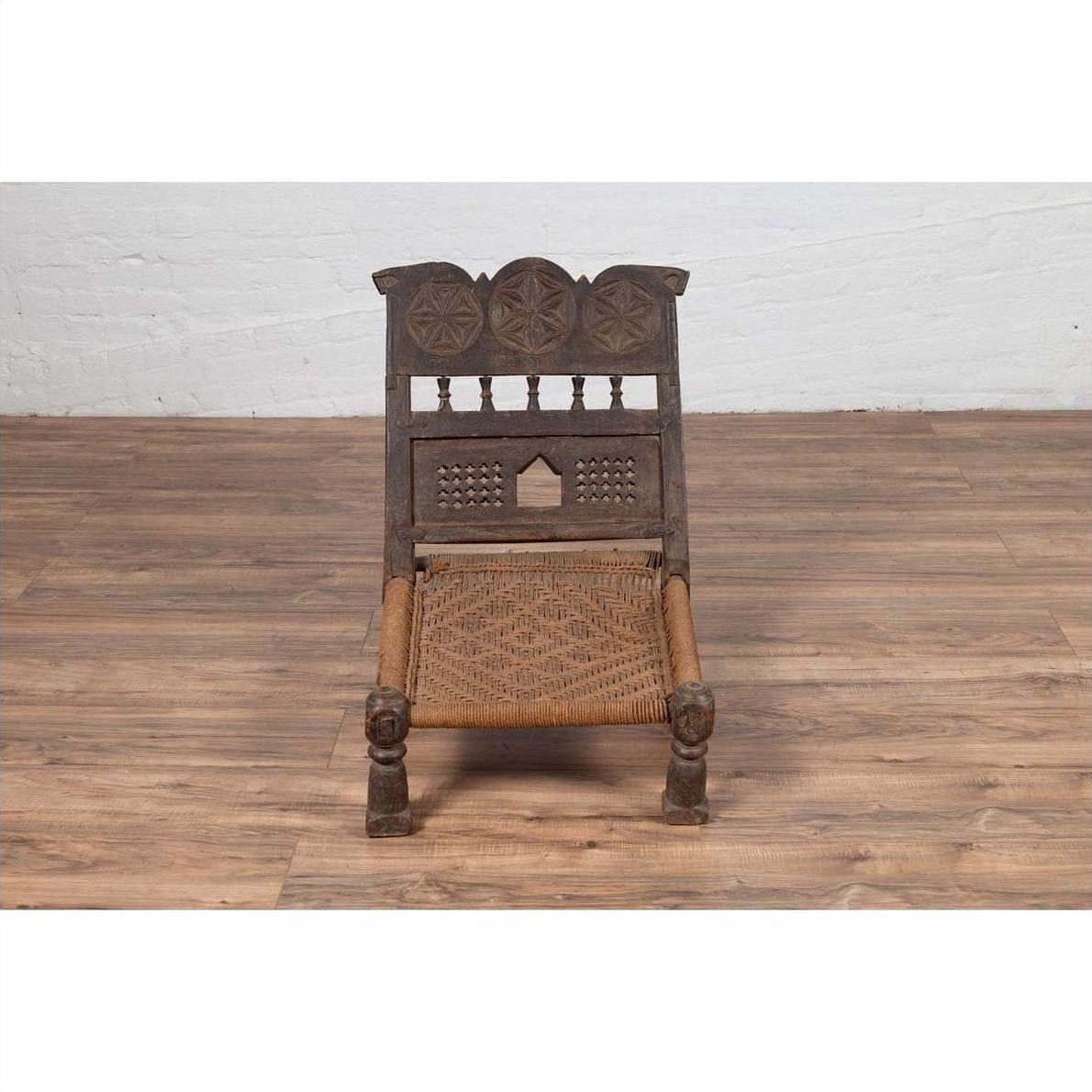 Brown wooden low seat chair with carved rosettes and handwoven rope seat, rustic Indian antique style.