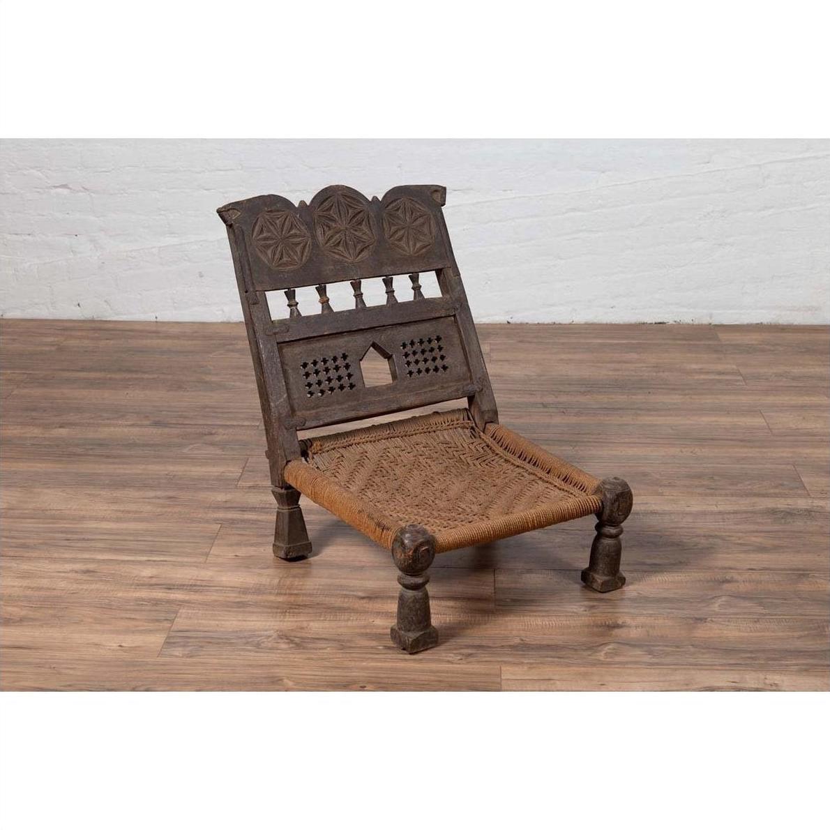 Brown wooden antique low seat chair with carved rosettes and rope seat on a wood floor.