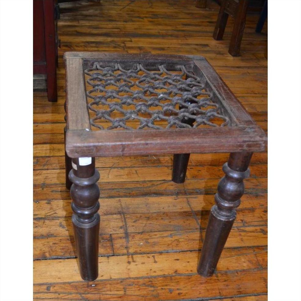 Brown wooden Indian side table with carved legs, lattice seat, and iron top on a wooden floor.