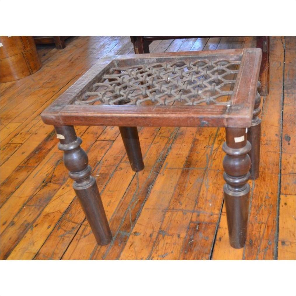 Dark wood Indian side table with ornate carving and four turned legs near window grill on polished wooden floor.