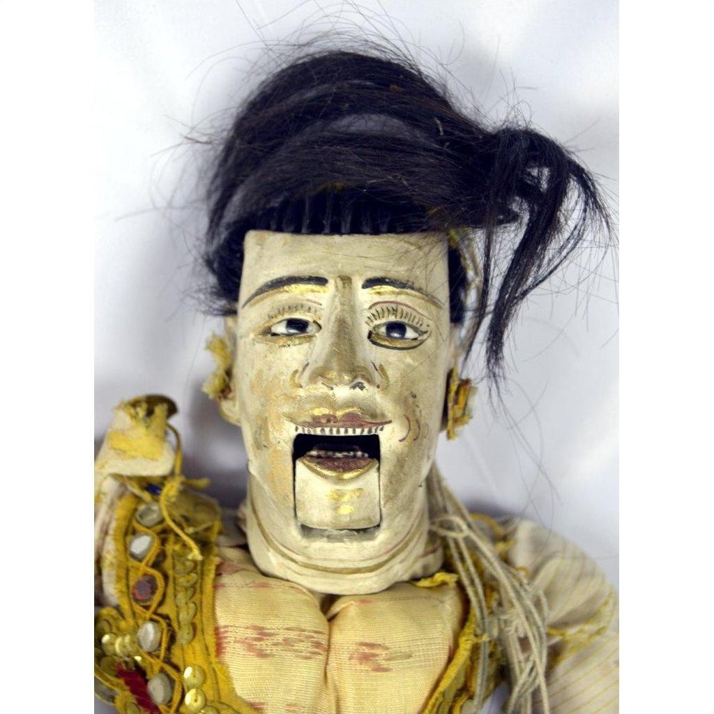 Close-up of a dark-haired Indonesian puppet with tangled strings and a moving mouth, set against a white background.