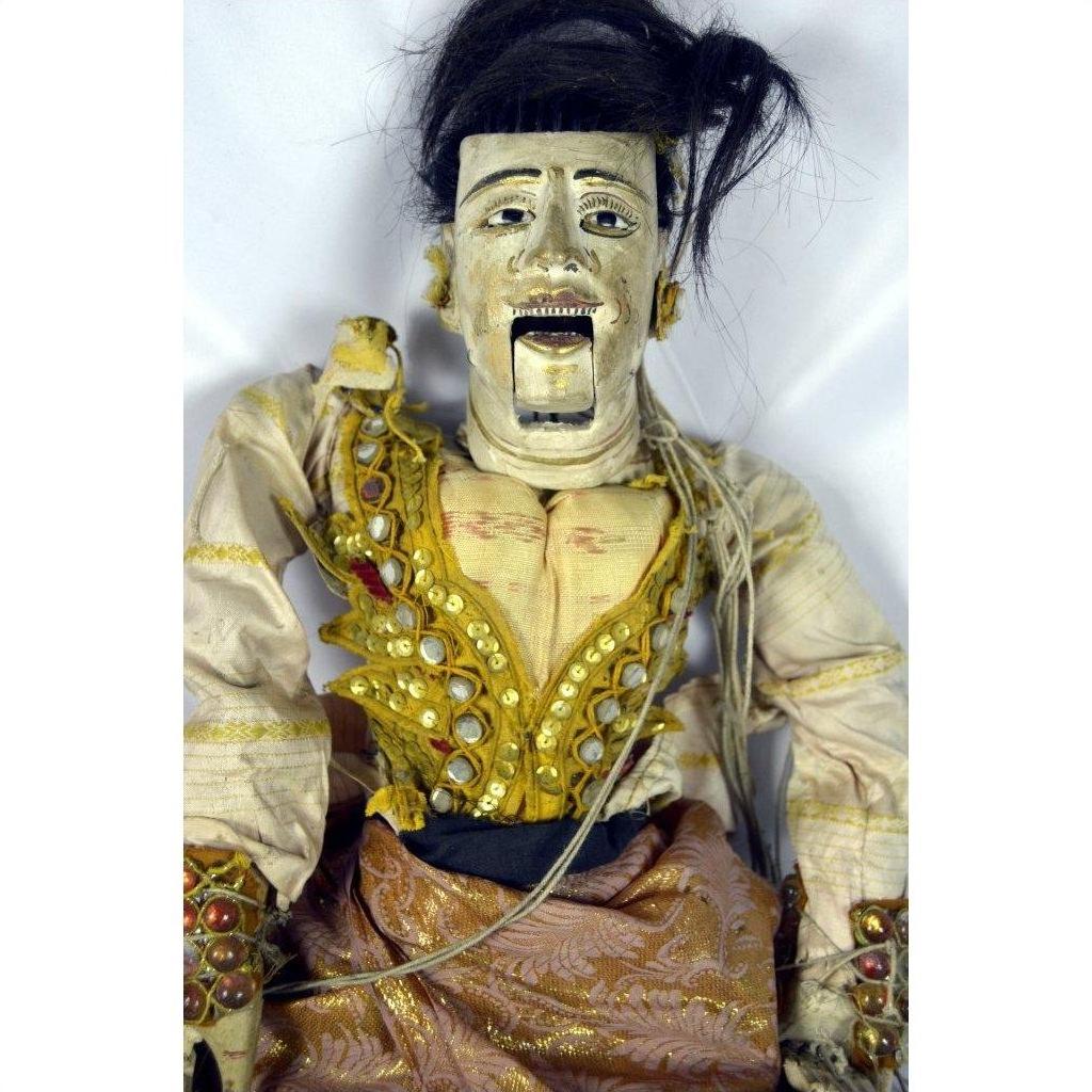 Close-up of Indonesian puppet with painted face, ornate jeweled clothing, black hair, and articulated fingers.