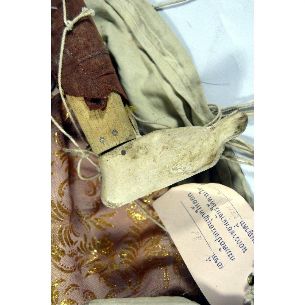 Brown wooden Indonesian puppet with white L-shaped end on patterned fabric, handwritten tag beside it.