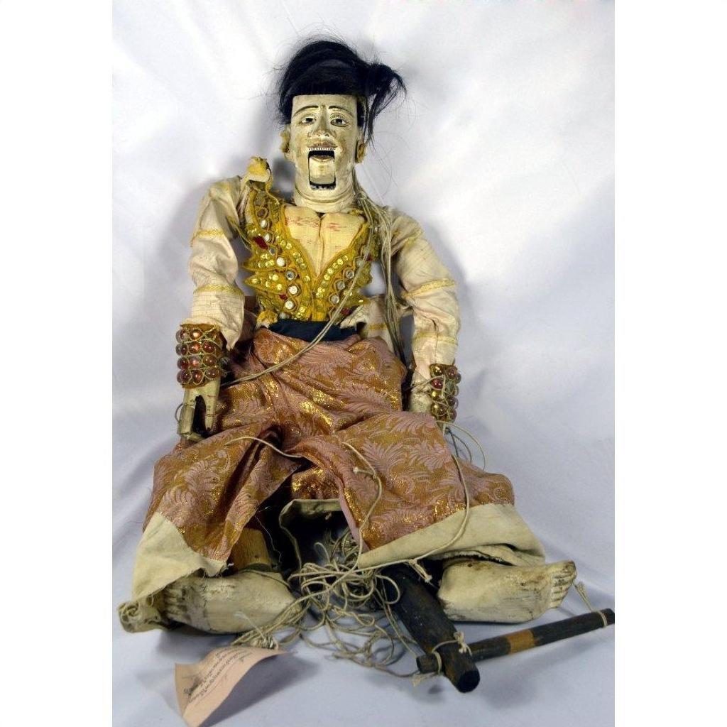 Brown wooden Indonesian puppet with detailed facial features, moving mouth, and articulated fingers, seated on plain backdrop.