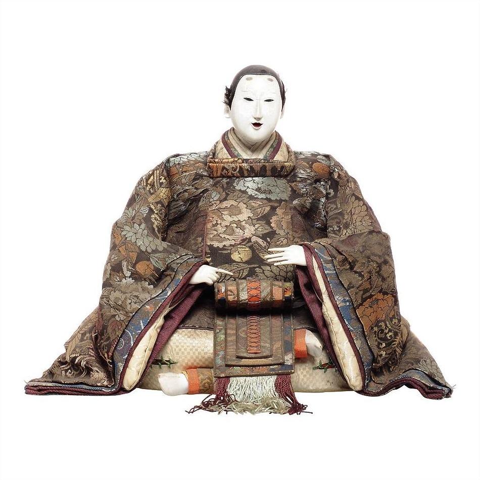 Close-up of Japanese doll in ornate Taisho Period kimono with white face and hands, seated against plain background.