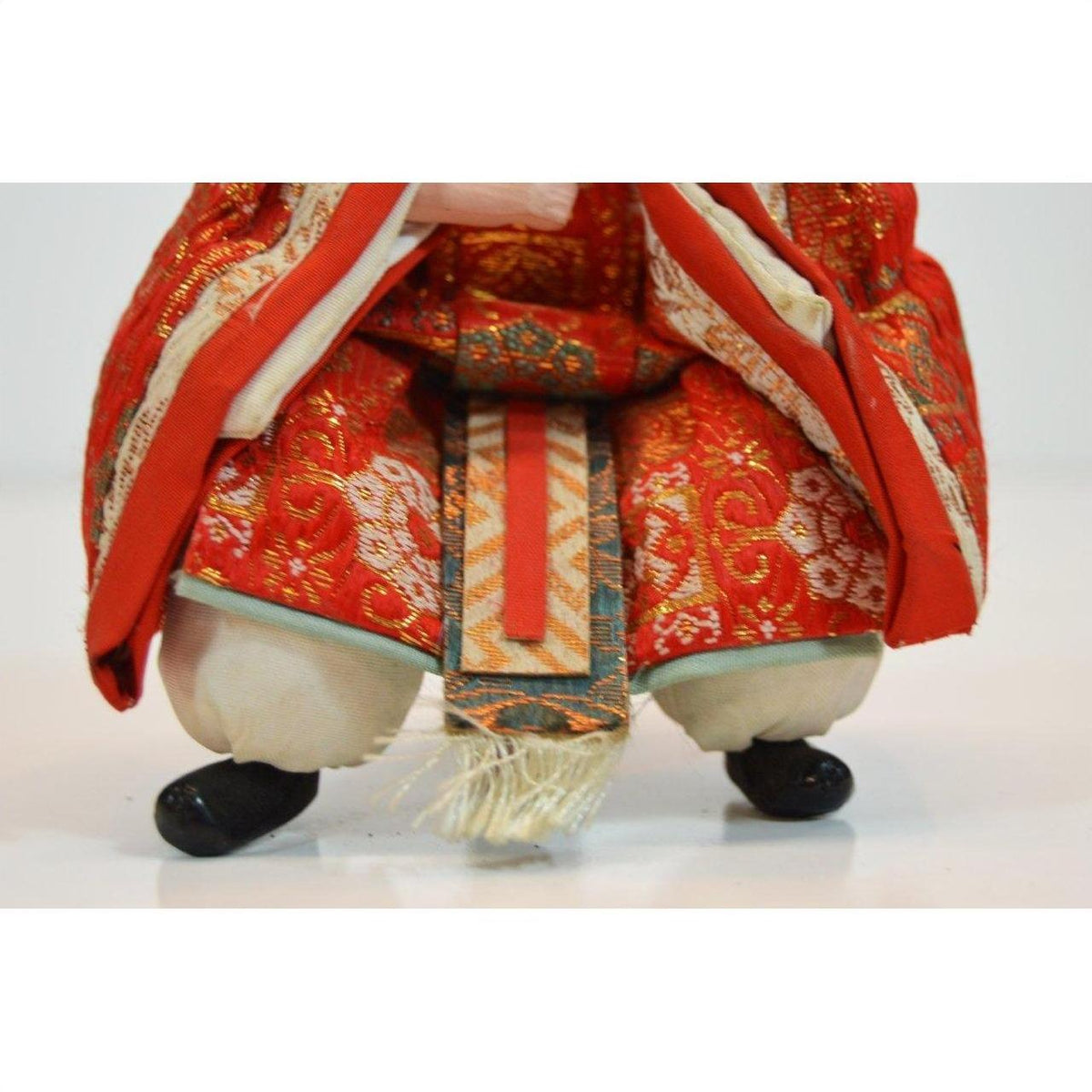 Close-up of Japanese doll in ornate red and gold fabric, kneeling with black shoes on white background.