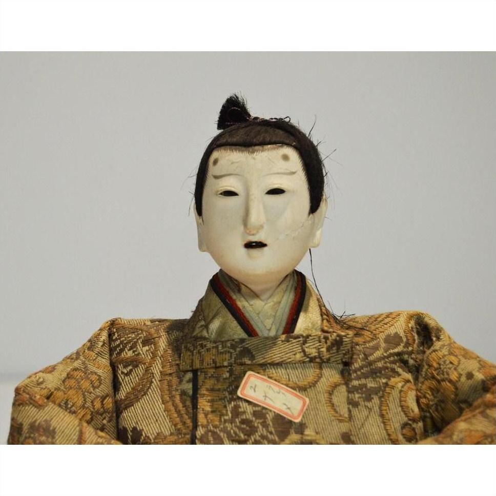 Close-up of Japanese Taisho samurai doll with white mask-like face and ornate patterned clothing, plain background.
