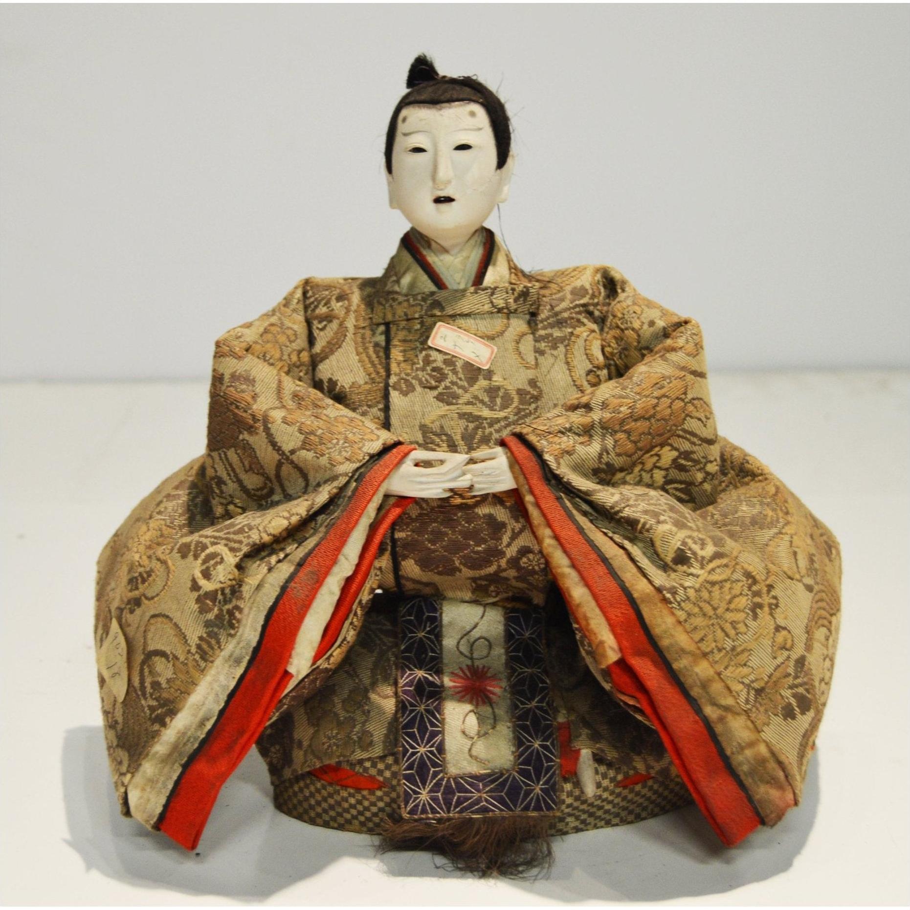 Ornate kimono Japanese Taisho Samurai Doll, folded arms, seated on plain surface, white background, close-up.
