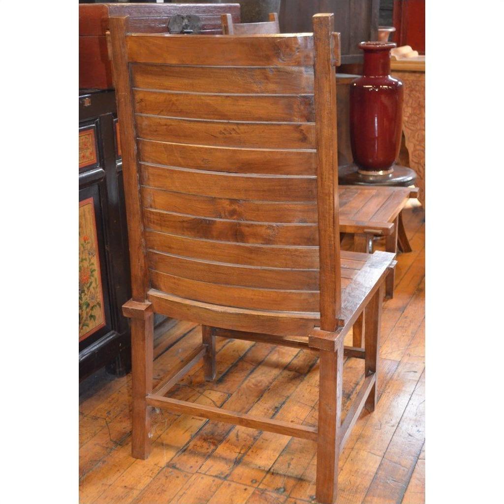 Brown teak wood side chair with slatted backrest on polished floor, furniture and red vase in background.