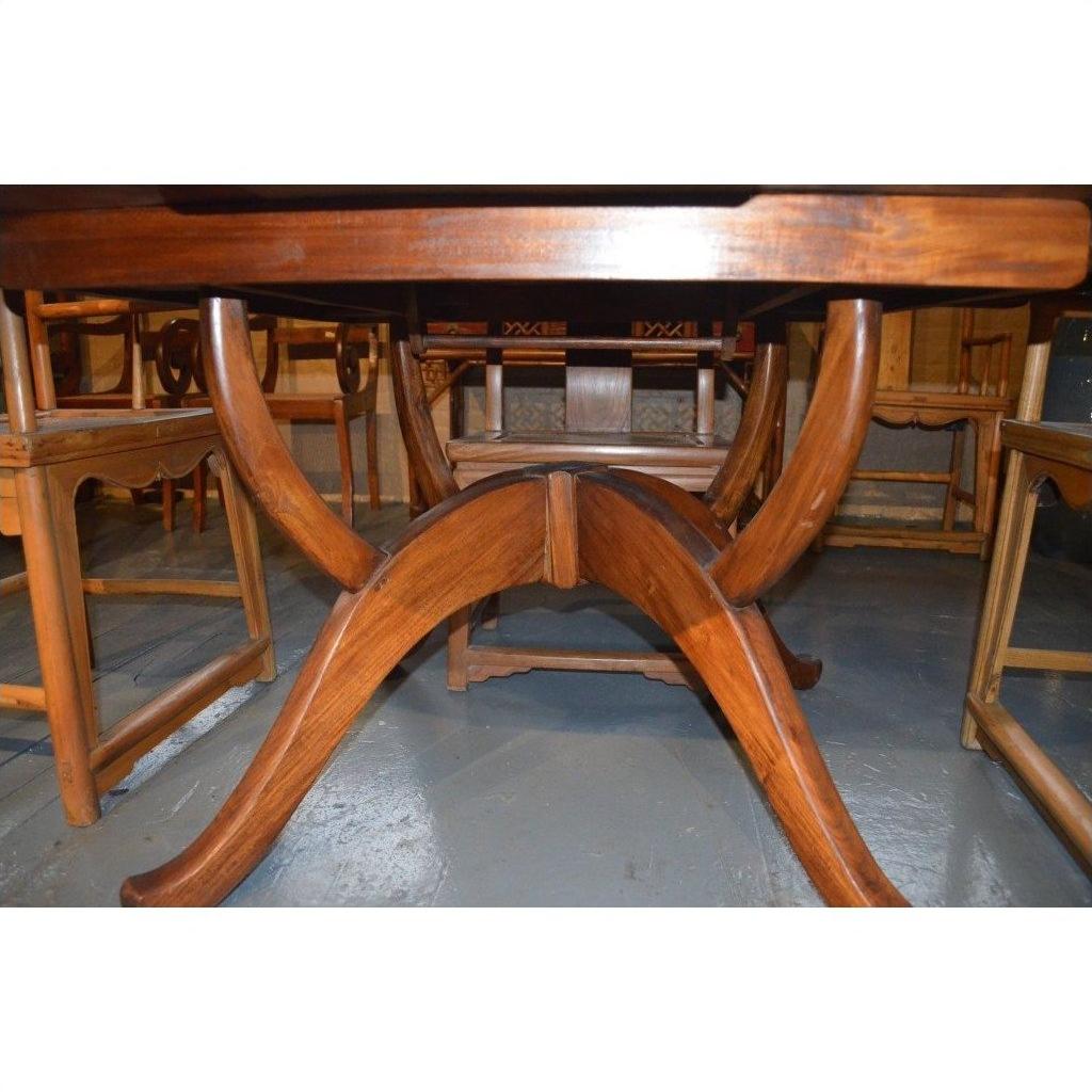 Brown wood Javanese oval dining table with leaf, viewed from below, surrounded by wooden chairs on a grey floor.