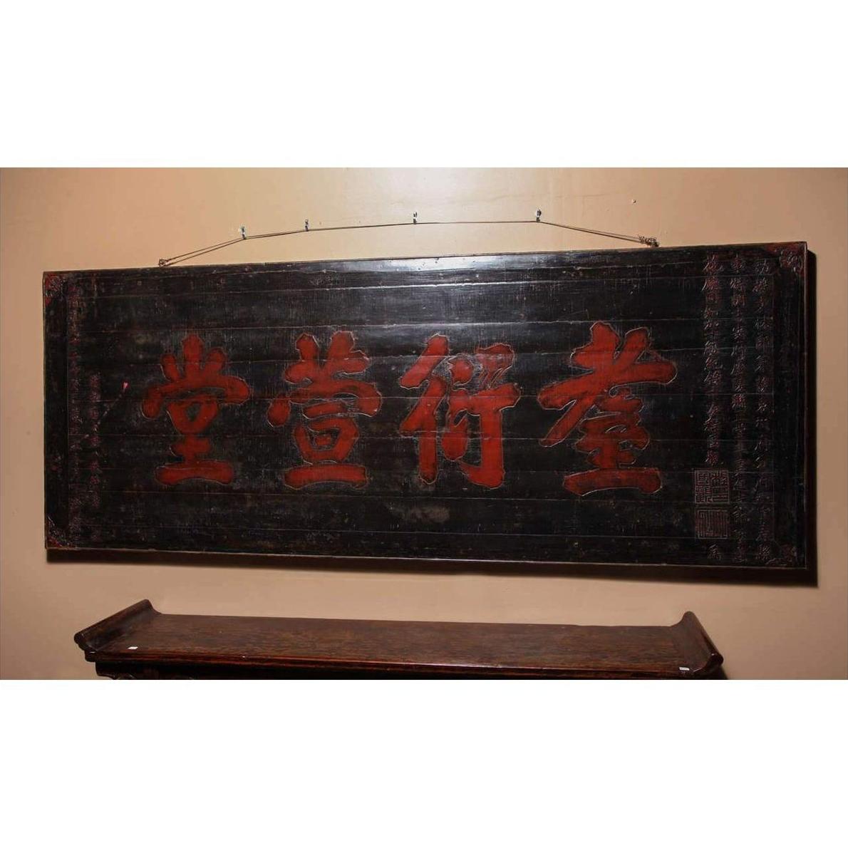 Red Chinese characters on 19th century wooden signboard, mounted above a wooden shelf on a beige wall.