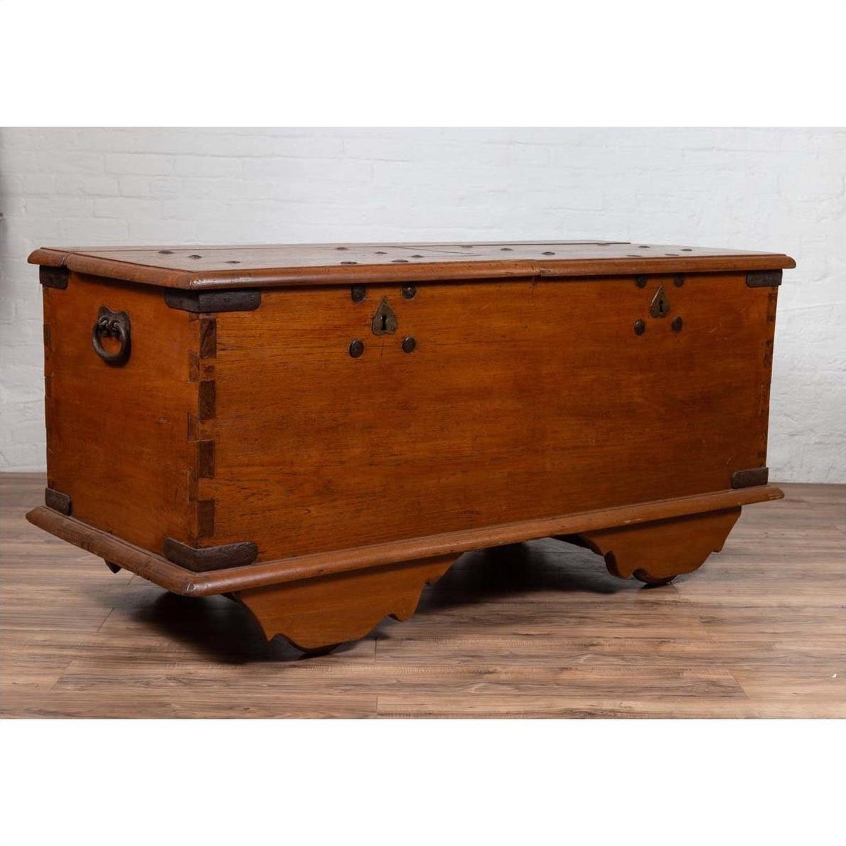 Brown teak wood chest with iron nailheads and wheels on a wooden floor next to a white brick wall indoors.
