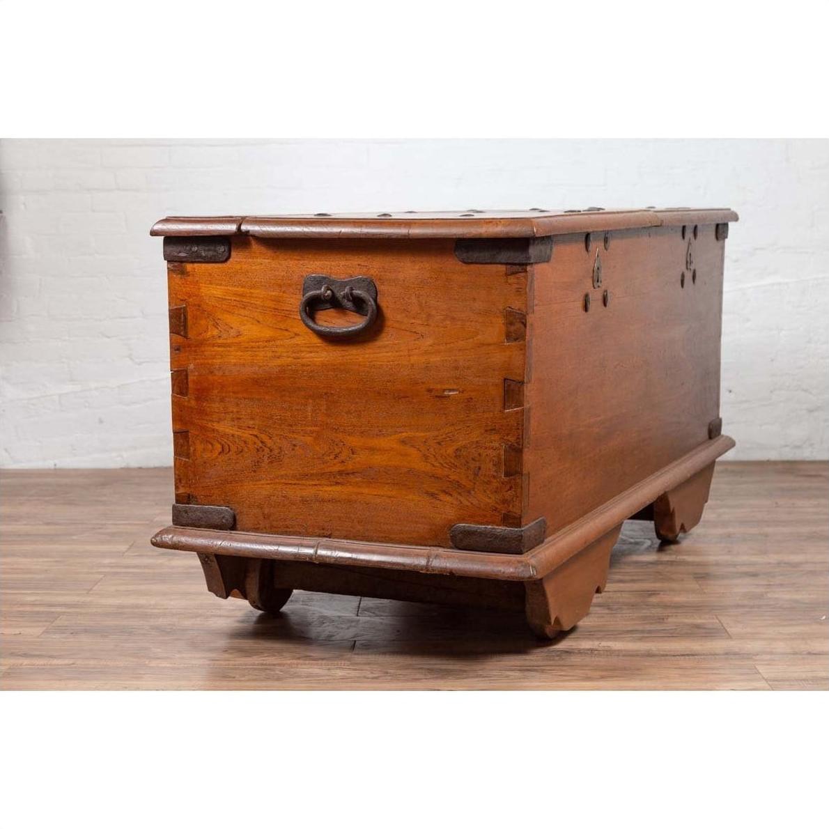 Brown teak wood blanket chest with iron nailheads on wheels, placed on a wooden floor by a white wall indoors.