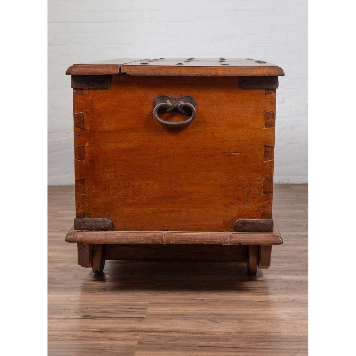 Brown teak wood chest with iron nailheads and wheels on wooden floor against a white brick wall indoors.