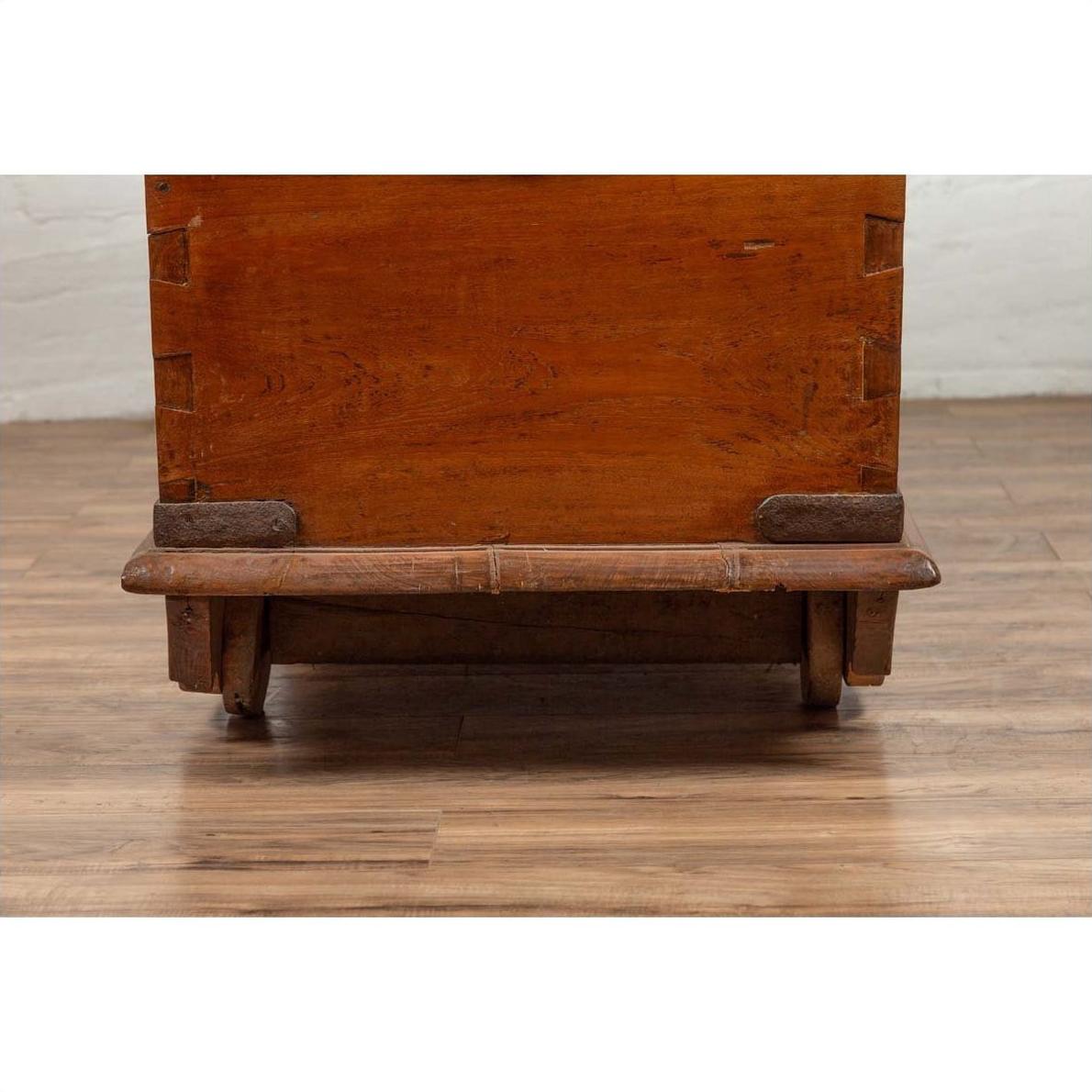 Close-up of brown teak wood blanket chest with iron nailheads on wheels, placed on a wood floor.