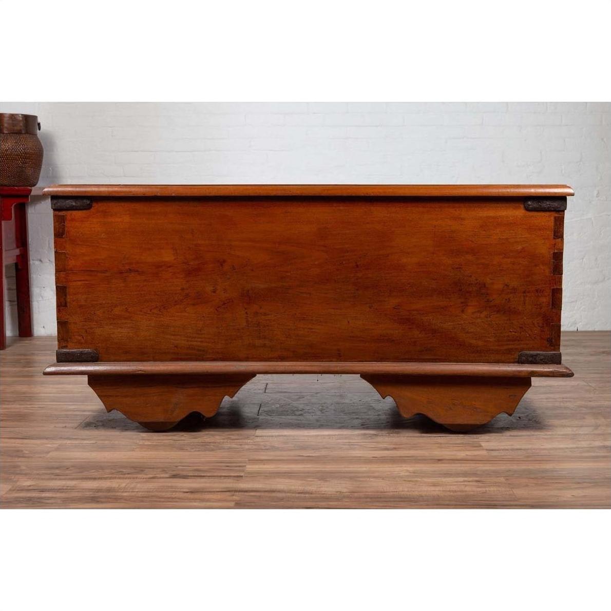 Brown teak wood chest with iron nailheads and wheels on a wooden floor, positioned against a white wall indoors.