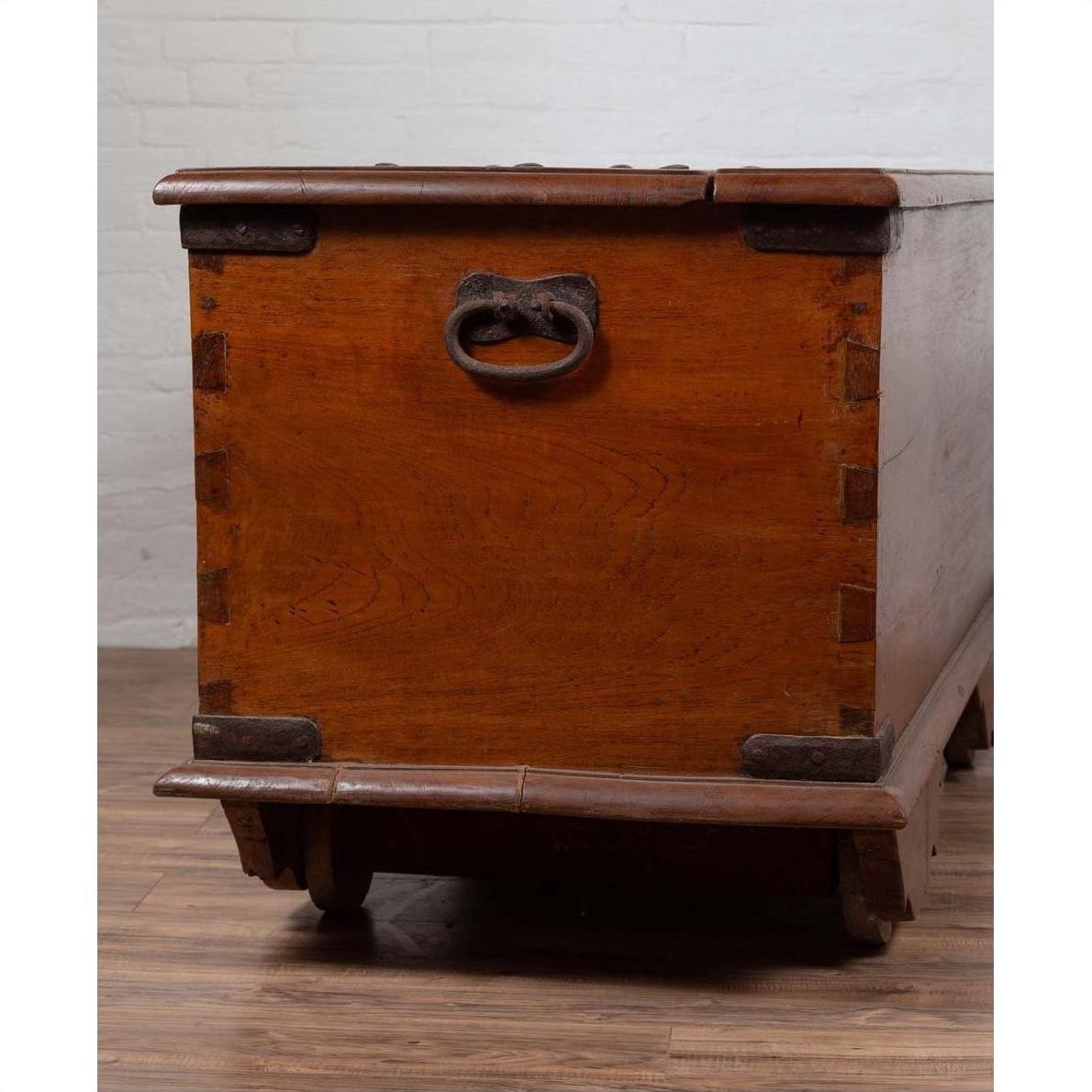 Brown teak wood chest with iron nailheads and wheels on a wooden floor against a white brick wall indoors.