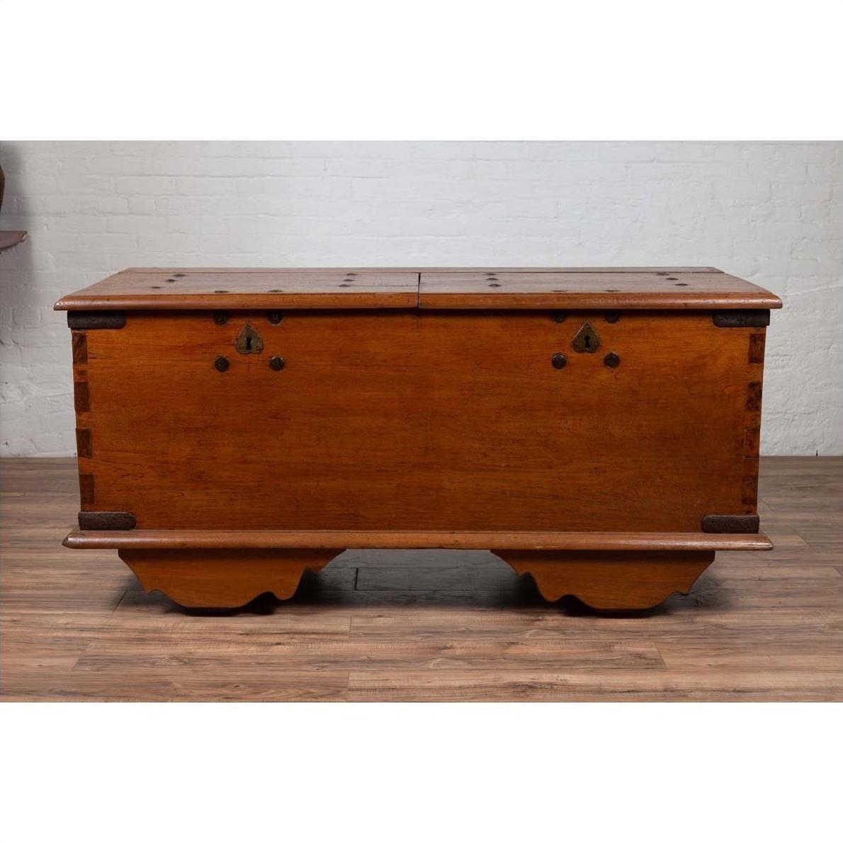 Brown teak wood chest with iron nailheads and wheels on wooden floor next to a white brick wall indoors.