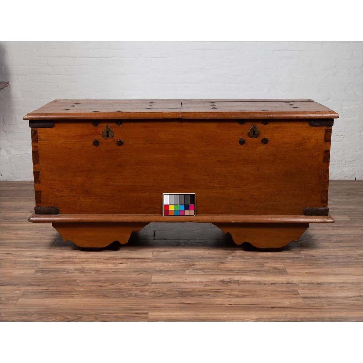 Brown teak wood blanket chest with iron nailheads on wheels, placed on wood floor next to a white brick wall indoors.