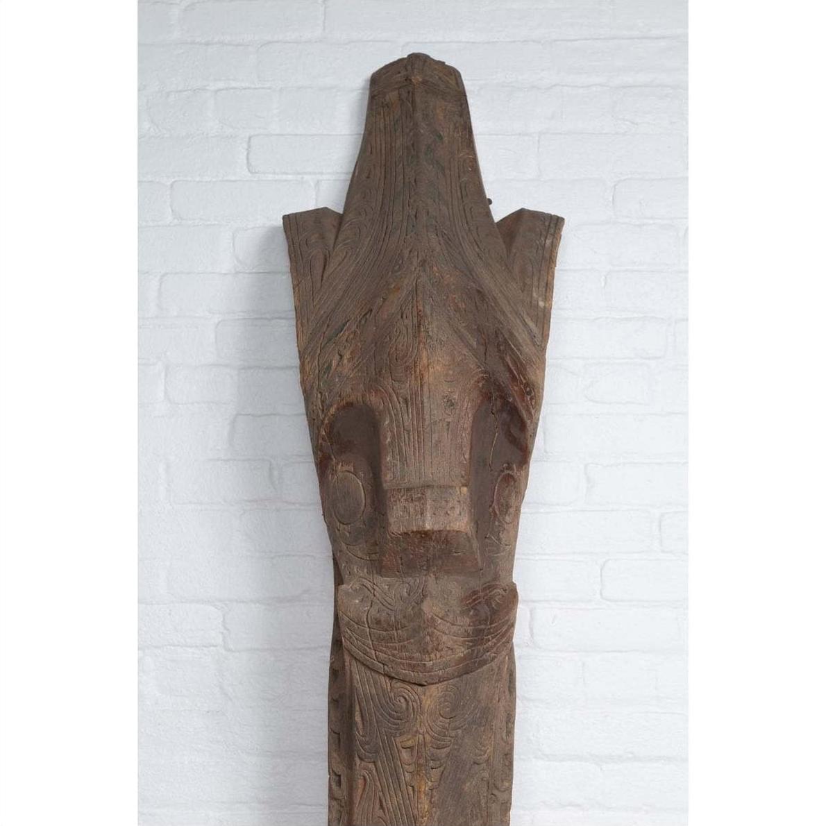 Brown wood Singa Singa Batak tribal face carving, hand carved, displayed against a white brick wall.