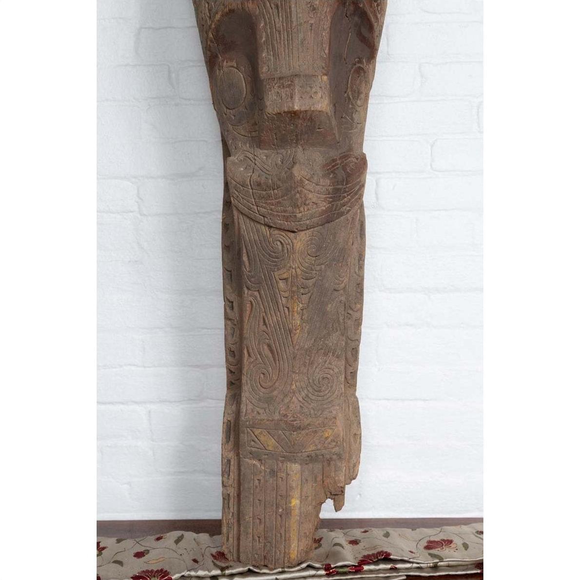 Brown hand-carved wooden Singa Singa tribal carving from Batak, Sumatra, against a white brick wall on patterned carpet.