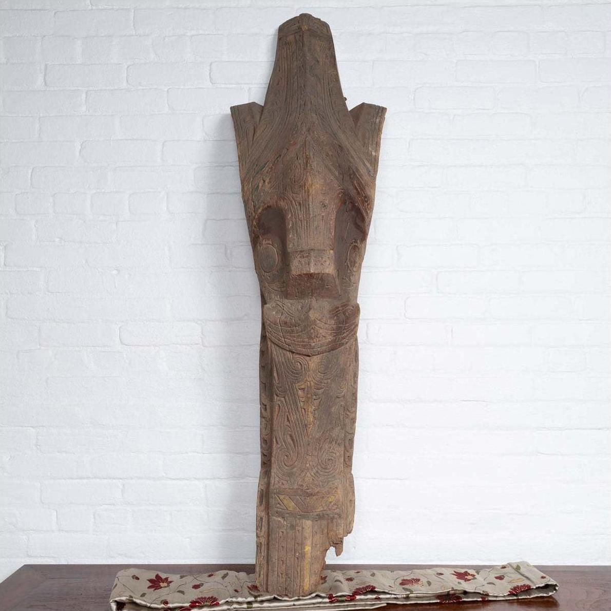 Brown wood Singa Singa tribal carving from the Batak people of Sumatra, on patterned cloth with brick wall background.