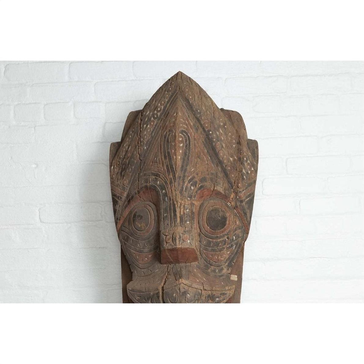 Brown hand carved wooden Singa Singa tribal mask from Batak people of Sumatra, displayed against white brick wall.