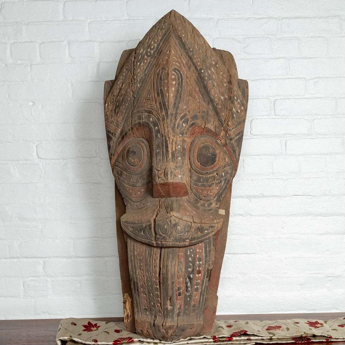 Brown wood Singa Singa tribal mask from Batak people, Sumatra, on patterned cloth against a white brick wall.