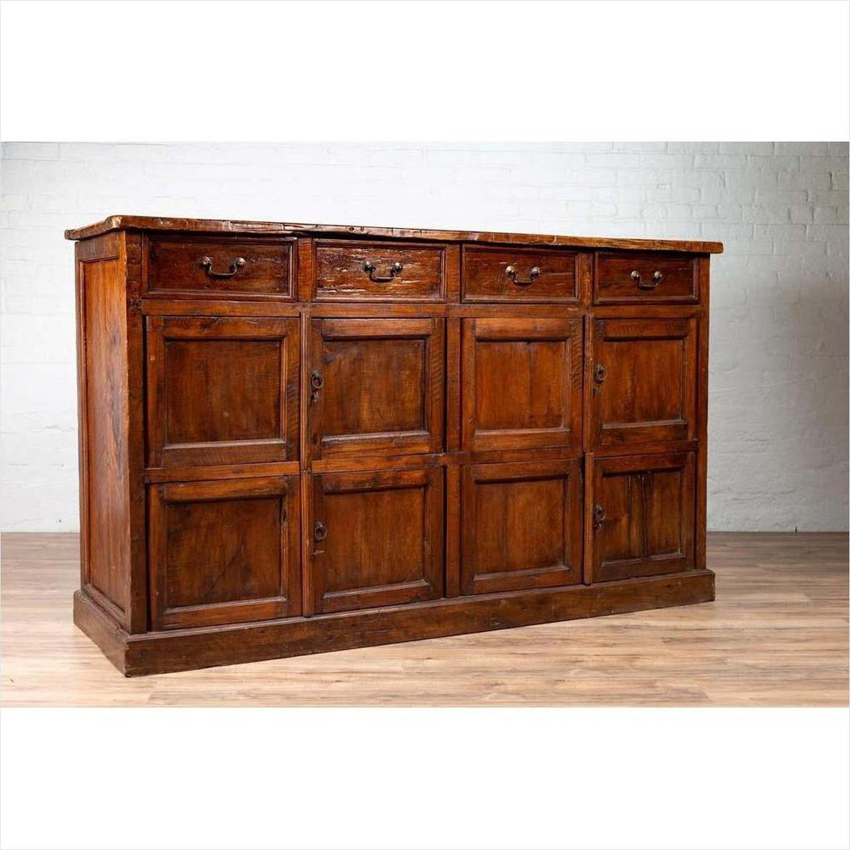 Brown teak wood cabinet with four drawers and four double doors, against white brick wall on wood floor indoors.