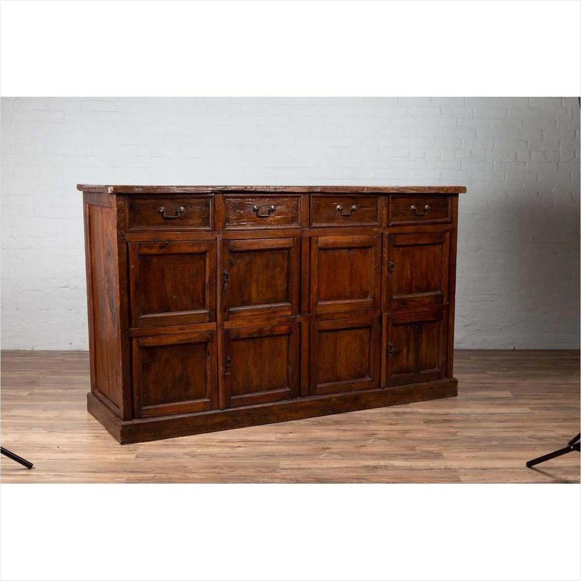 Brown teak wood cabinet with four drawers and four double doors, set against a white brick wall indoors.