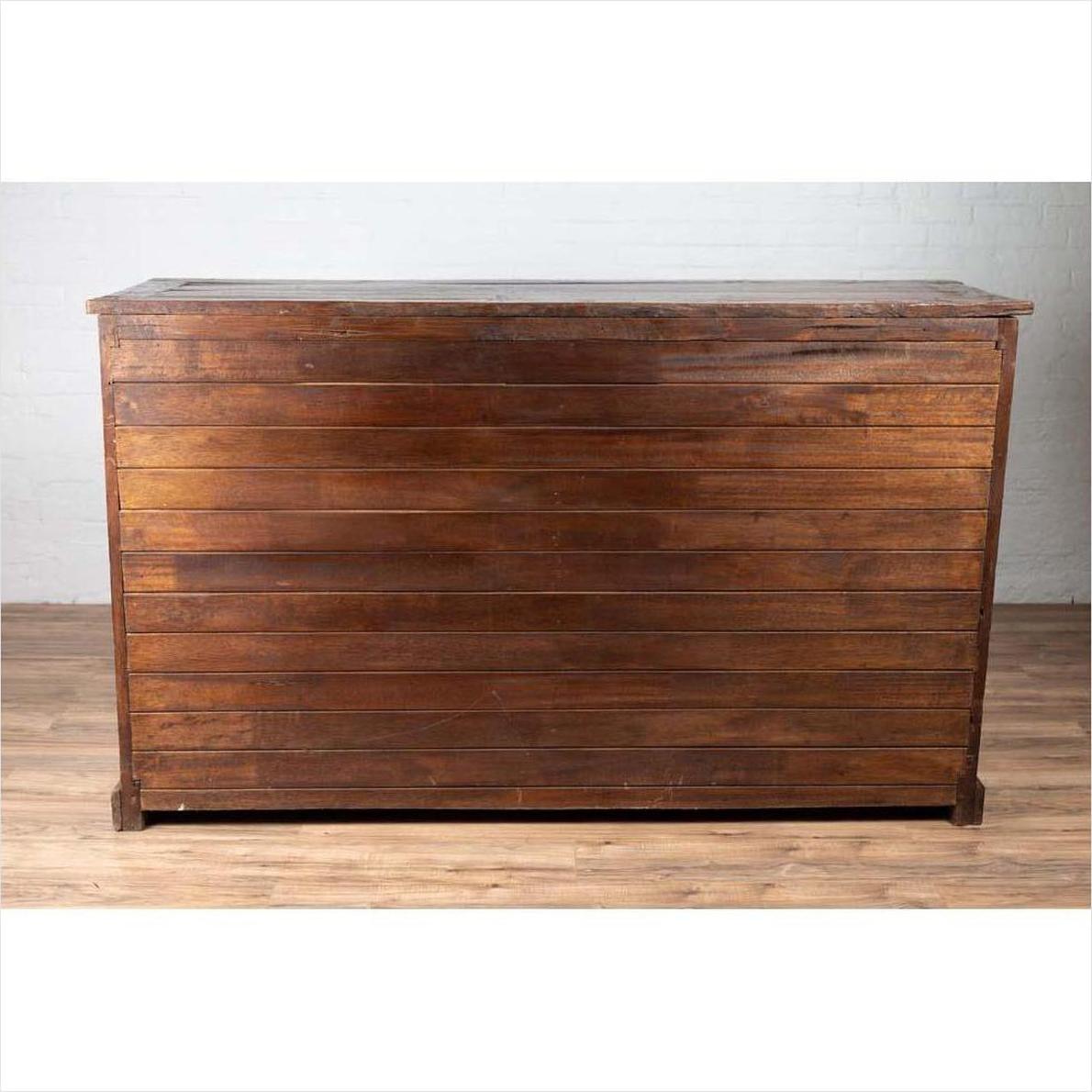 Brown teak wood cabinet with four drawers and double doors, placed on a wooden floor against a white brick wall indoors.