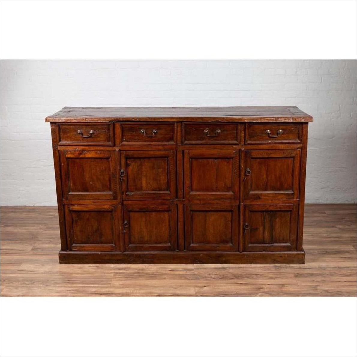Brown teak wood cabinet with four drawers and four double doors, placed indoors against a white brick wall.